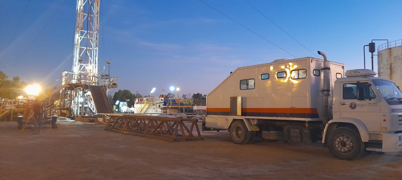 Unidad móvil de Slickline junto a torre de perforación al atardecer